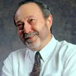 Smiling man in a white shirt and patterned tie against a dark background.