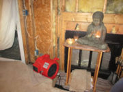 A Flood of Blessings Buddha statue on a wooden table next to a red blower in an unfinished wooden room.