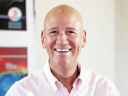 Organic Intelligence Smiling bald man in a white shirt in a bright room.