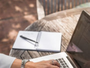 Volume 7 Number 3 Winter 2018 Person typing on a laptop at a wooden table with a notebook and pen.