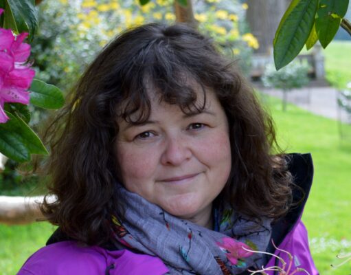 In the Bleak Midwinter Smiling woman in a purple jacket among green plants with pink flowers.